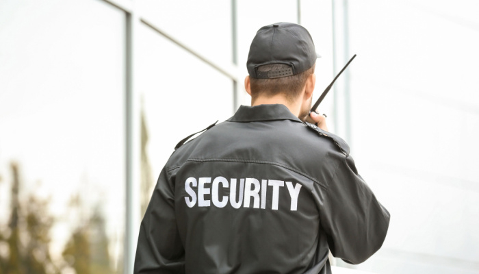 Male security guard in black cap using portable radio transmitter near building outdoors communicating with someone